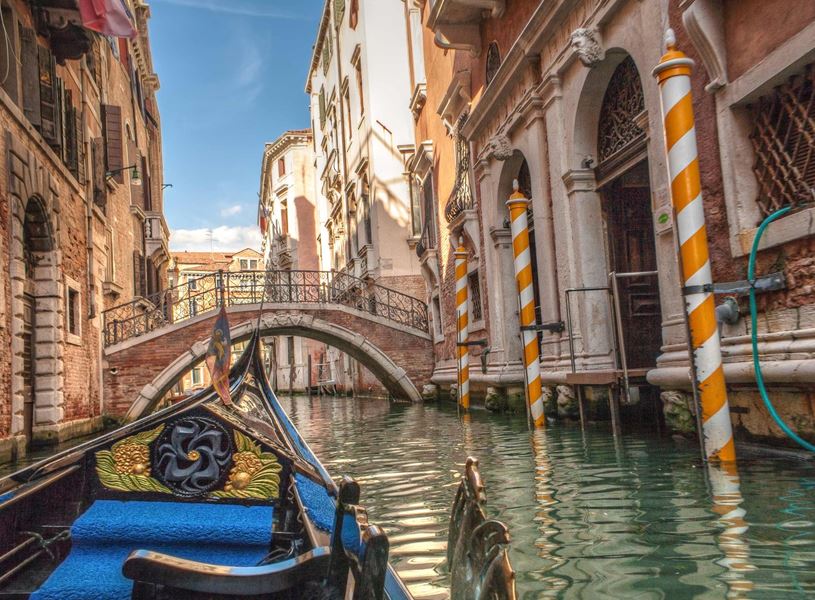 Gondola gliding through Venetian canal toward arched bridge with striped mooring poles 