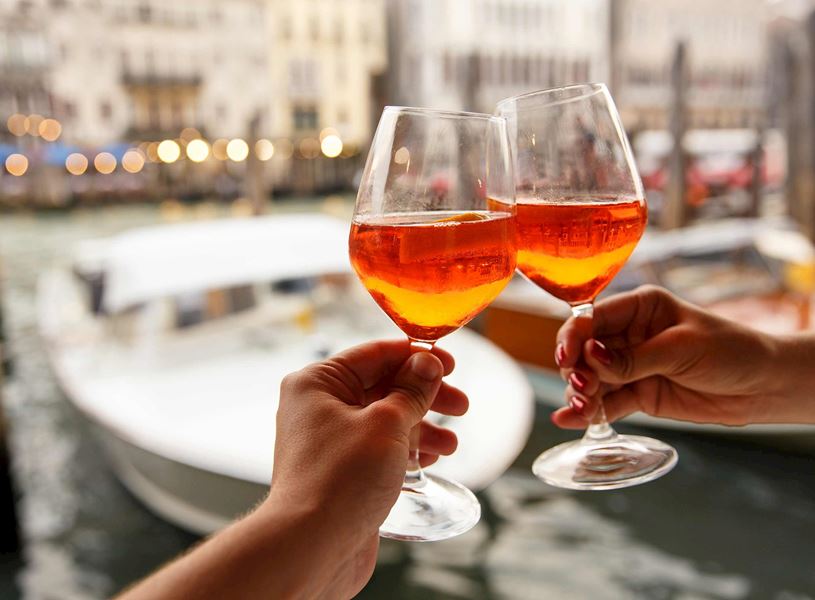 Two hands clinking glasses of Aperol Spritz with blurred boats in background