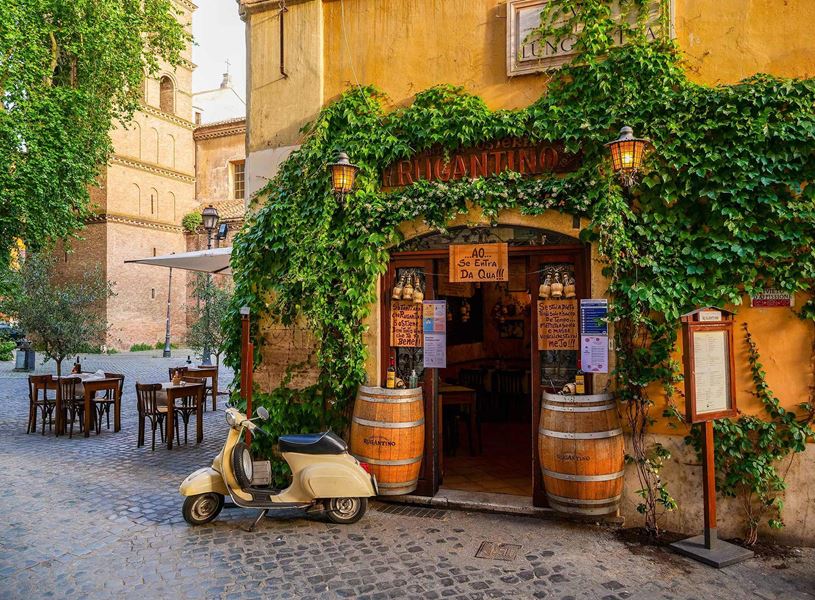 Vintage Vespa parked outside ivy-covered Italian restaurant in Rome with wooden barrels and tables.