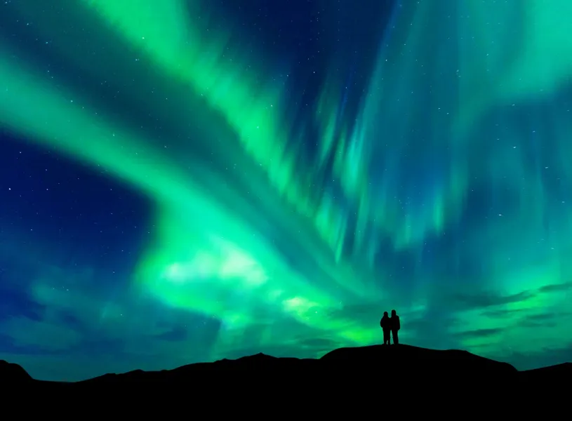 Northern Lights over hilltop with two silhouetted figures observing in Reykjavik, Iceland