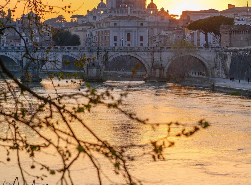 St. Peter’s Basilica at sunset viewed across Tiber River with bridge and tree branches in foreground