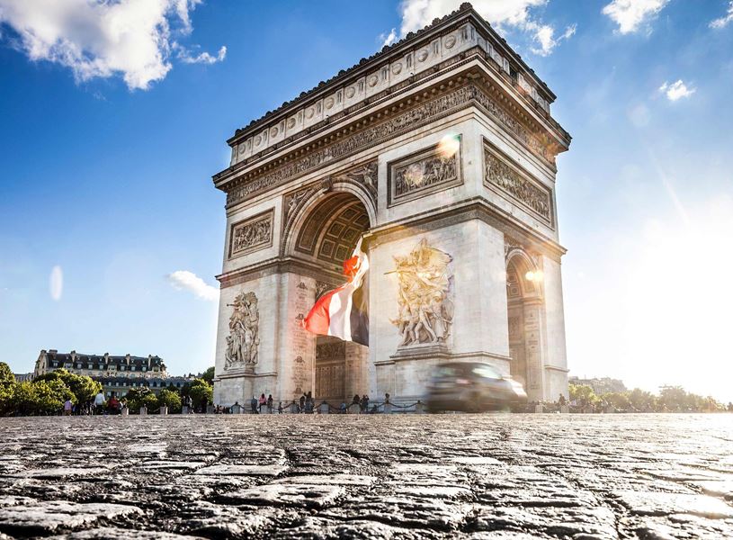 Arc de Triomphe with French flag, sun flare and cobblestone foreground in Paris, France