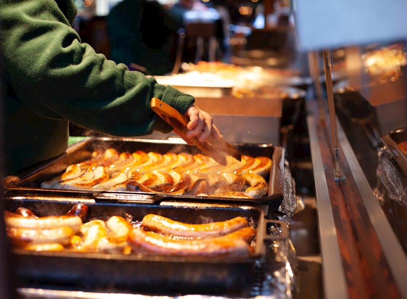 Hands using tongs to grill bratwurst sausages at food stall.