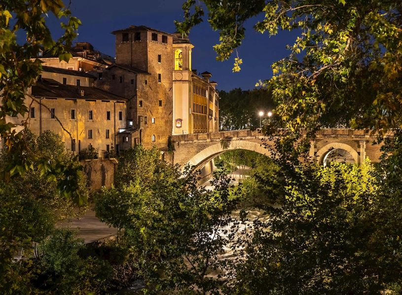 Night view of Tiber Island in Rome with illuminated stone bridge and historic buildings framed by trees