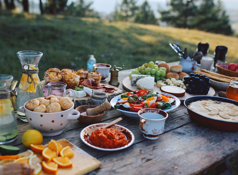 Rustic outdoor table filled with assorted fresh fruits, cheeses, bread, salads and drinks
