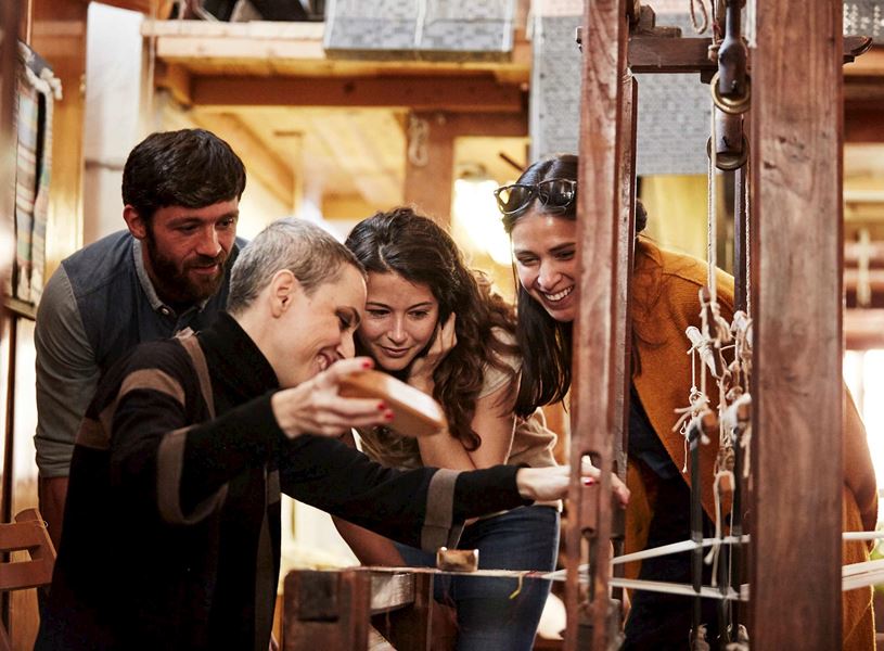 Group observing traditional wooden loom weaving process inside rustic workshop setting