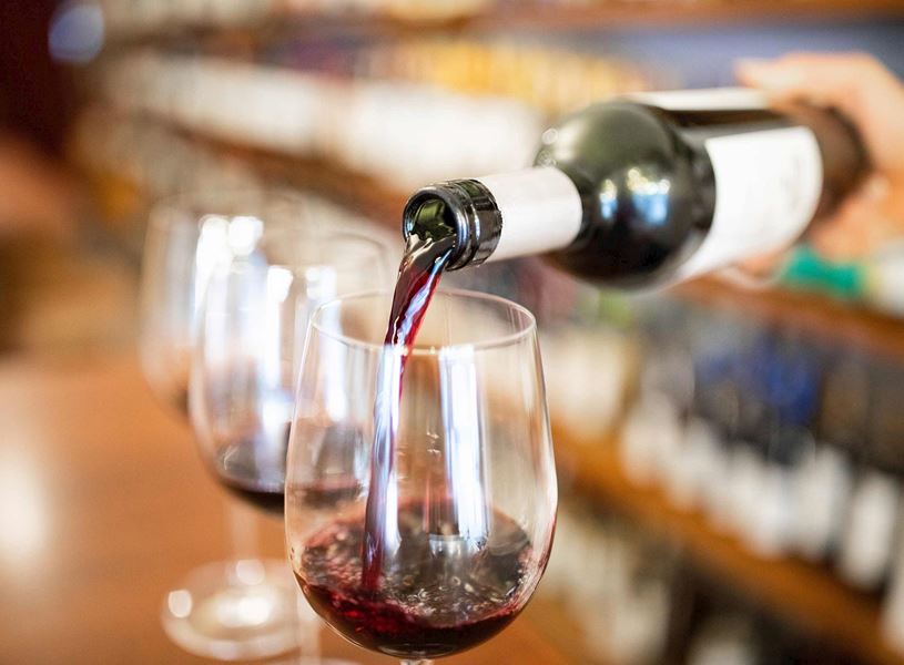Red wine being poured from bottle into glass on wooden counter with blurred wine shelves