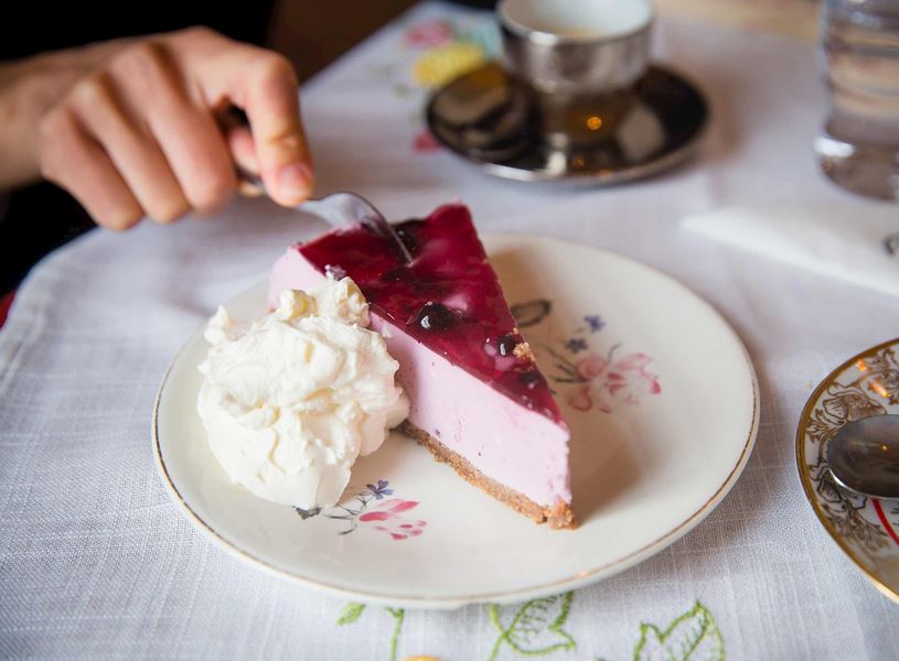 Hand holding fork over berry cheesecake slice with whipped cream on plate