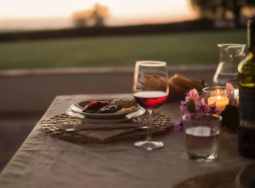 Outdoor dining table with glass of red wine, bread, candlelight and flowers at sunset