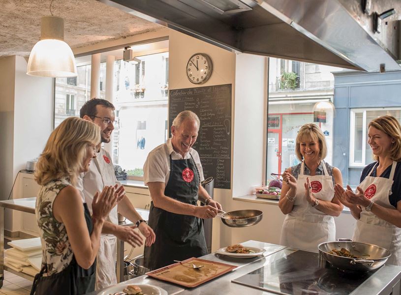 Cooking class group in aprons gathered around stove in kitchen.
