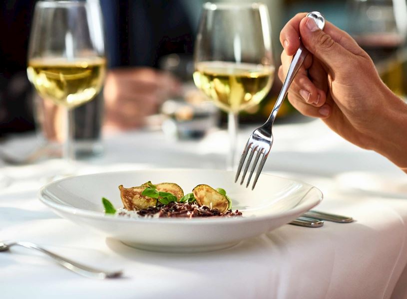 Hand holding fork over gourmet dish with wine glasses on table