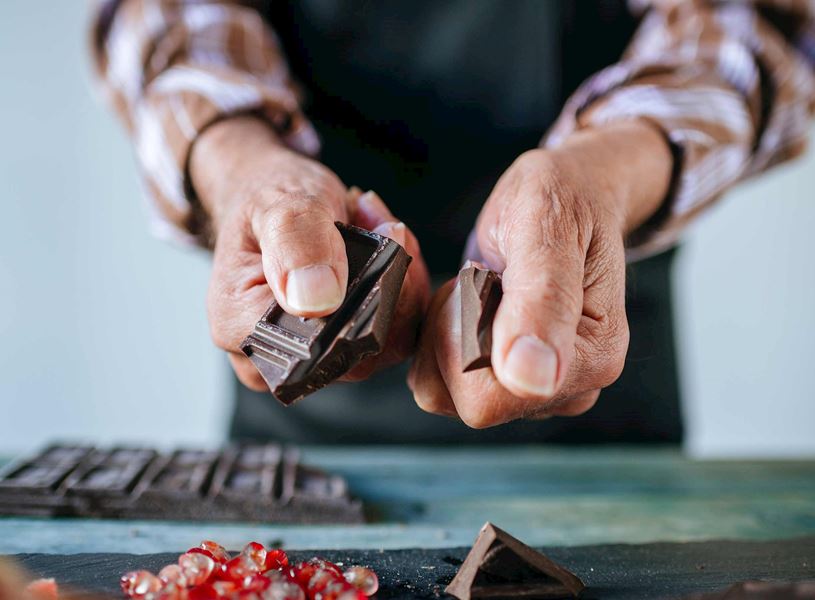 Hands breaking dark chocolate pieces on table with pomegranate seeds nearby