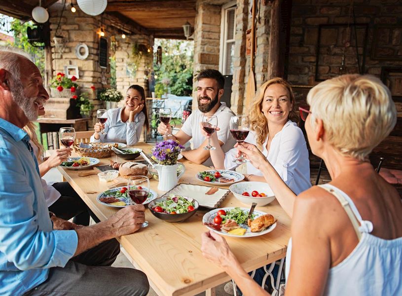 Group dining outdoors at wooden table with plates of salad, bread and glasses of red wine