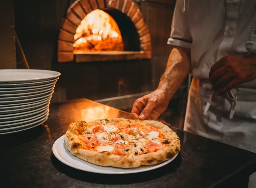 Freshly baked pizza on white plate placed by chef near wood-fired brick oven