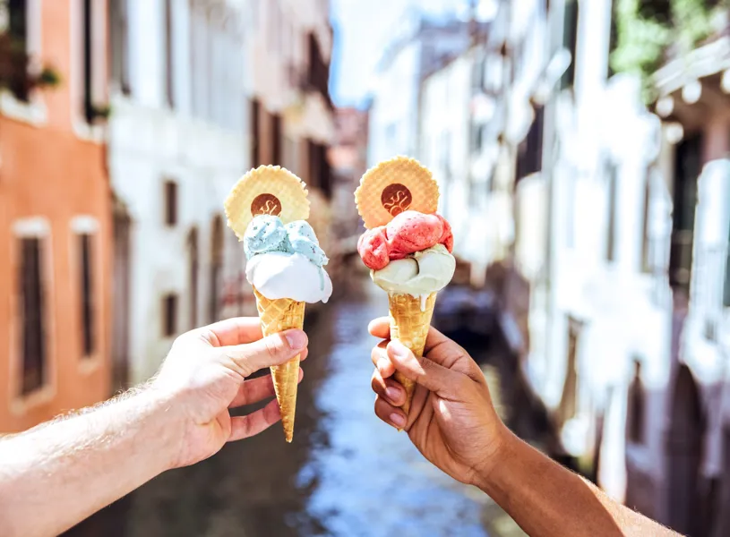 Two hands holding waffle cones with colourful gelato scoops and wafer discs above Venetian canal