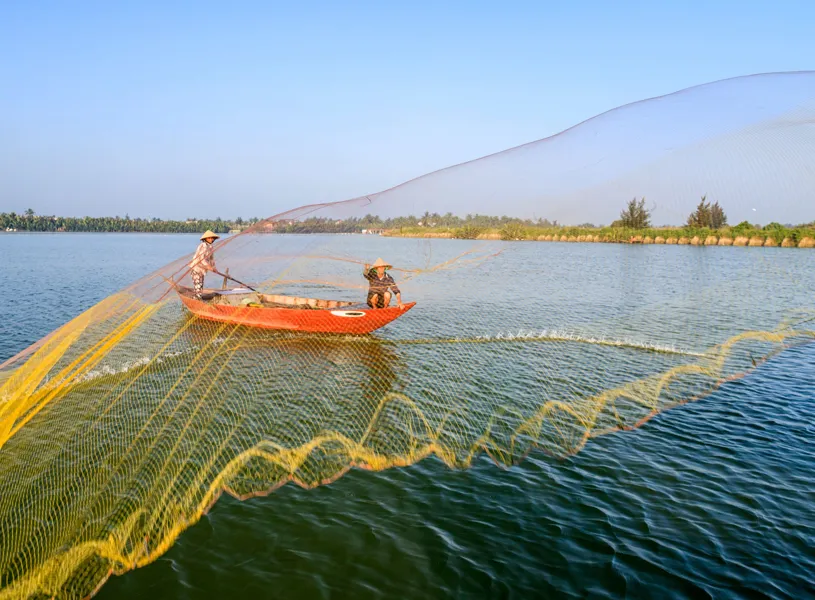 Fisher casting net from wooden boat on tranquil river Cua Dai, Hoi An, Vietnam