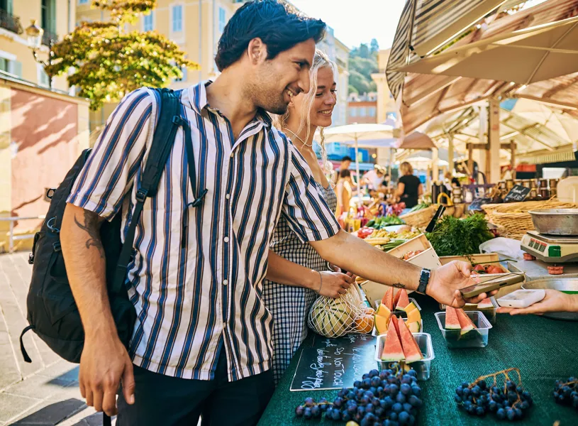 Two travellers buying fruit at a market stand, one using smartphone to pay