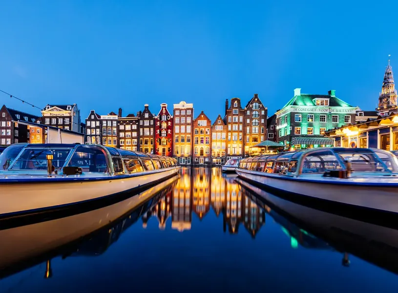Canal boats docked with illuminated traditional houses at dusk in Amsterdam, Netherlands
