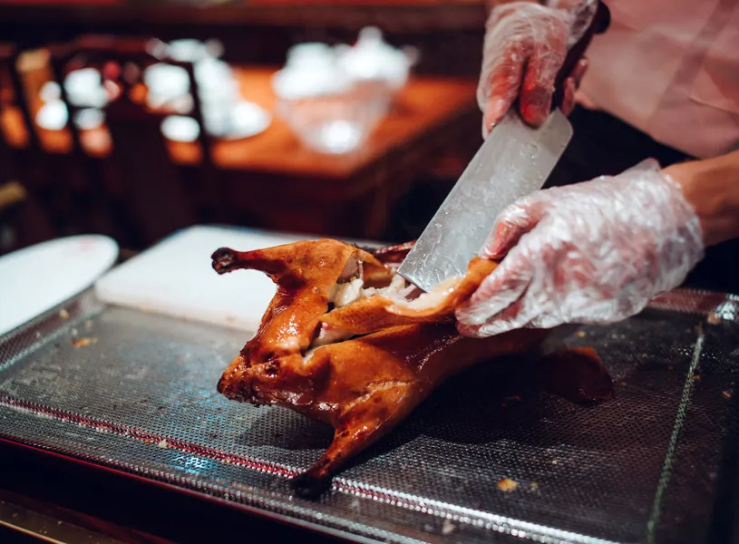 Gloved hands slicing whole roasted Peking duck with cleaver on tray, China