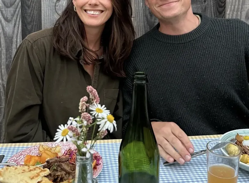 Male and female sitting at a table facing the camera with plates of food and drink infant of them.